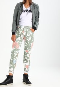 Světle zelená bomber bunda přes bílé tričko s černým textem, v kombinaci s vzorovanými slim-fit kalhotami s růžovými a modrými květinovými motivy a černými botami.