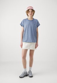 Tricou din bumbac de culoare albastru deschis, cu mâneci răsfrânte, purtat cu o fustă din denim albă cu buzunare în față. Persoana poartă adidași gri și o pălărie cu dungi.