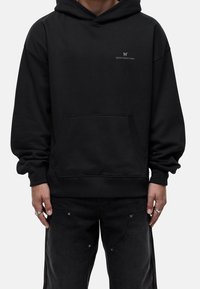 Persona con una sudadera negra con bolsillo frontal y un pequeño logotipo "GOODFORNOTHING" en el pecho, combinada con pantalones negros y anillos plateados.