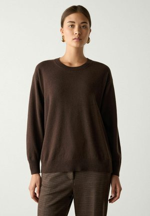 Ung kvinde iført en mørkebrun langærmet sweater, ternede brune bukser og guld øreringe i ringeform står mod en ensfarvet baggrund.