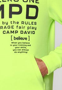 Felpa con cappuccio verde neon caratterizzata da audaci motivi in nero, una tasca frontale e una consistenza morbida. Il testo include frasi motivazionali e dettagli del marchio.