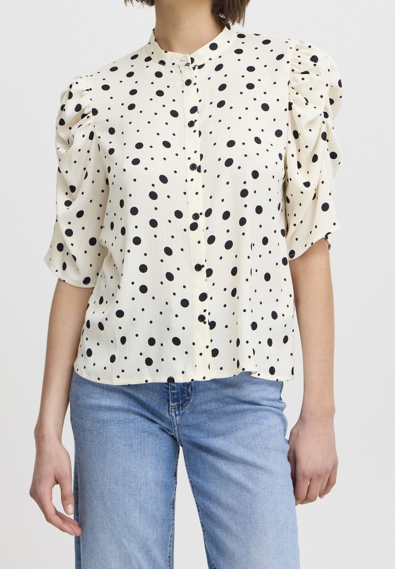 Short-sleeve blouse in cream fabric, featuring black polka dots, a buttoned front, and gathered sleeves, paired with light blue jeans.