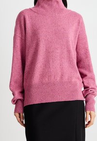 Pull en laine rose avec col camionneur, doté de poignets et d'un ourlet côtelés, offrant une texture douce et une coupe décontractée, associé à une jupe noire.