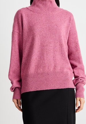 Pull en laine rose avec col camionneur, doté de poignets et d'un ourlet côtelés, offrant une texture douce et une coupe décontractée, associé à une jupe noire.