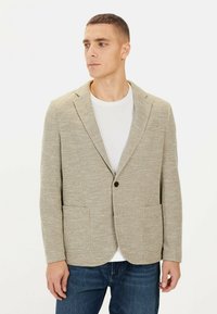 Helles beige Blazer aus strukturiertem Stoff, mit Reverskragen, einreihiger Knopfschluss und zwei Vorderentaschen, getragen über einem weißen T-Shirt.