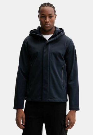 Uomo con capelli intrecciati che indossa una giacca nera con cappuccio e tasche con cerniera, in piedi davanti a uno sfondo bianco.