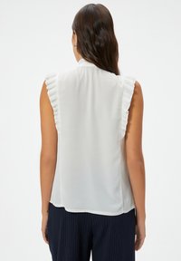 Blusa blanca sin mangas con acentos de hombros con volantes, tela suave y un corte holgado. Llevada con pantalones de raya diplomática en azul marino, vista desde atrás.