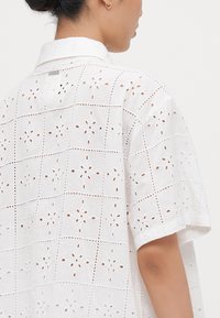 Camisa de algodón blanca con bordado de ojo de flor en un patrón de cuadrícula, mangas cortas y cuello puntiagudo, con un ajuste relajado.