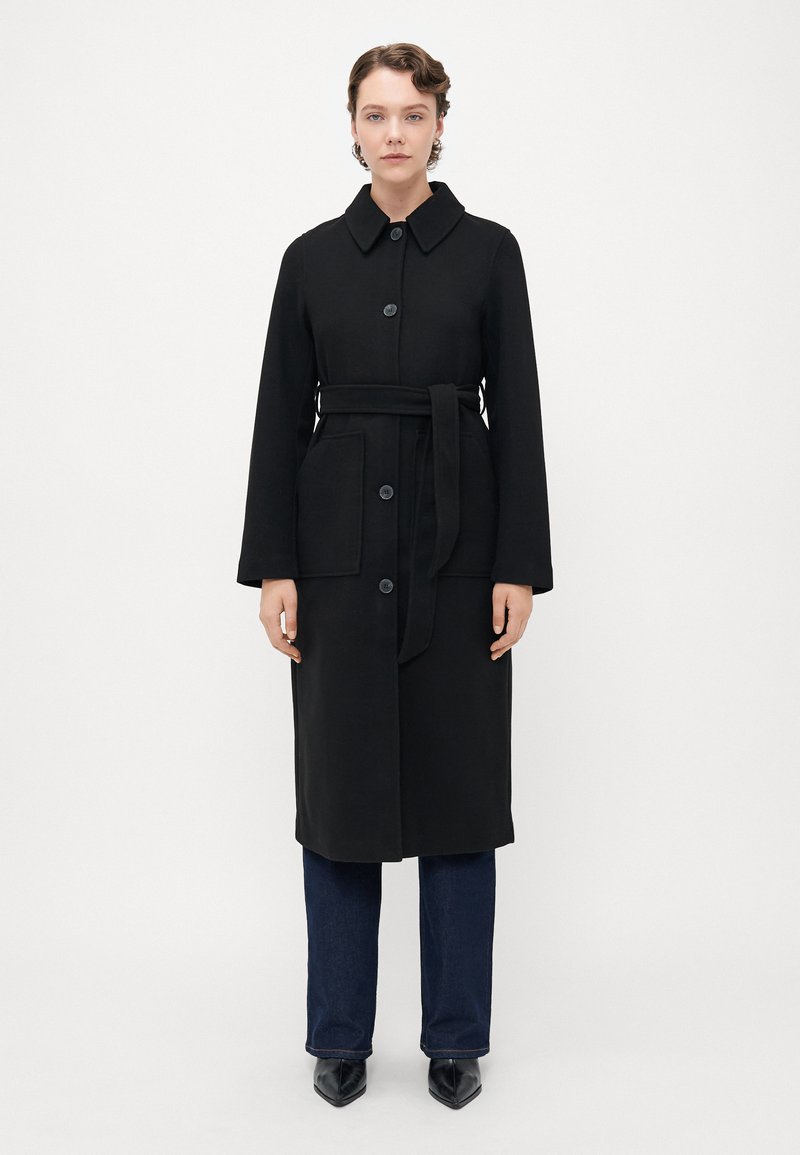 Donna in piedi davanti a uno sfondo bianco, indossa un lungo cappotto nero abbottonato con cintura, jeans blu scuro e scarpe nere a punta.