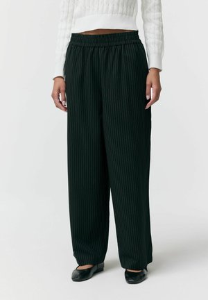 Osoba ubraną w czarne, szerokie spodnie w prążki, biały teksturowany sweter i czarne baleriny, stojącą na jednolitym tle.