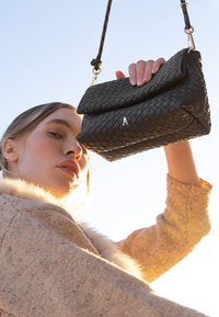 Sac à main en cuir noir tressé avec un rabat souple rabattable, agrémenté d’un logo en métal argenté et d’une sangle à motif. Design compact et texturé.