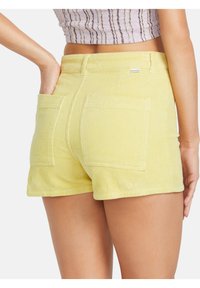 Keltaiset corduroy-shortsit, joissa on tekstuuripinta, kaksi takataskua ja rento malli, näkyvät takaa.