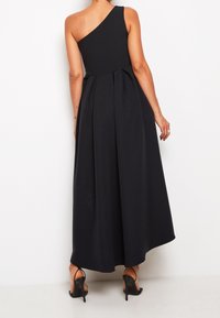 Robe longue noire asymétrique avec un dos plissé et une silhouette fluide. Tissu à la texture lisse. Portée avec des talons noirs à brides.