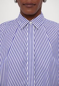 Blå och vit randig skjorta med spetsig krage, med vertikala ränder och knäppning framtill. Slätt tyg med en strukturerad design.