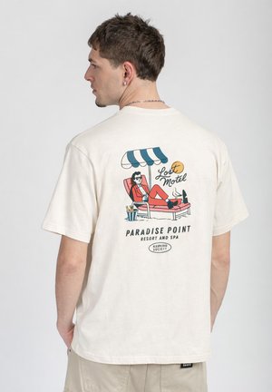 Man wearing white t-shirt with colorful graphic of person lounging under teal-and-white umbrella, text "Lost Motel Paradise Point Resort and Spa."