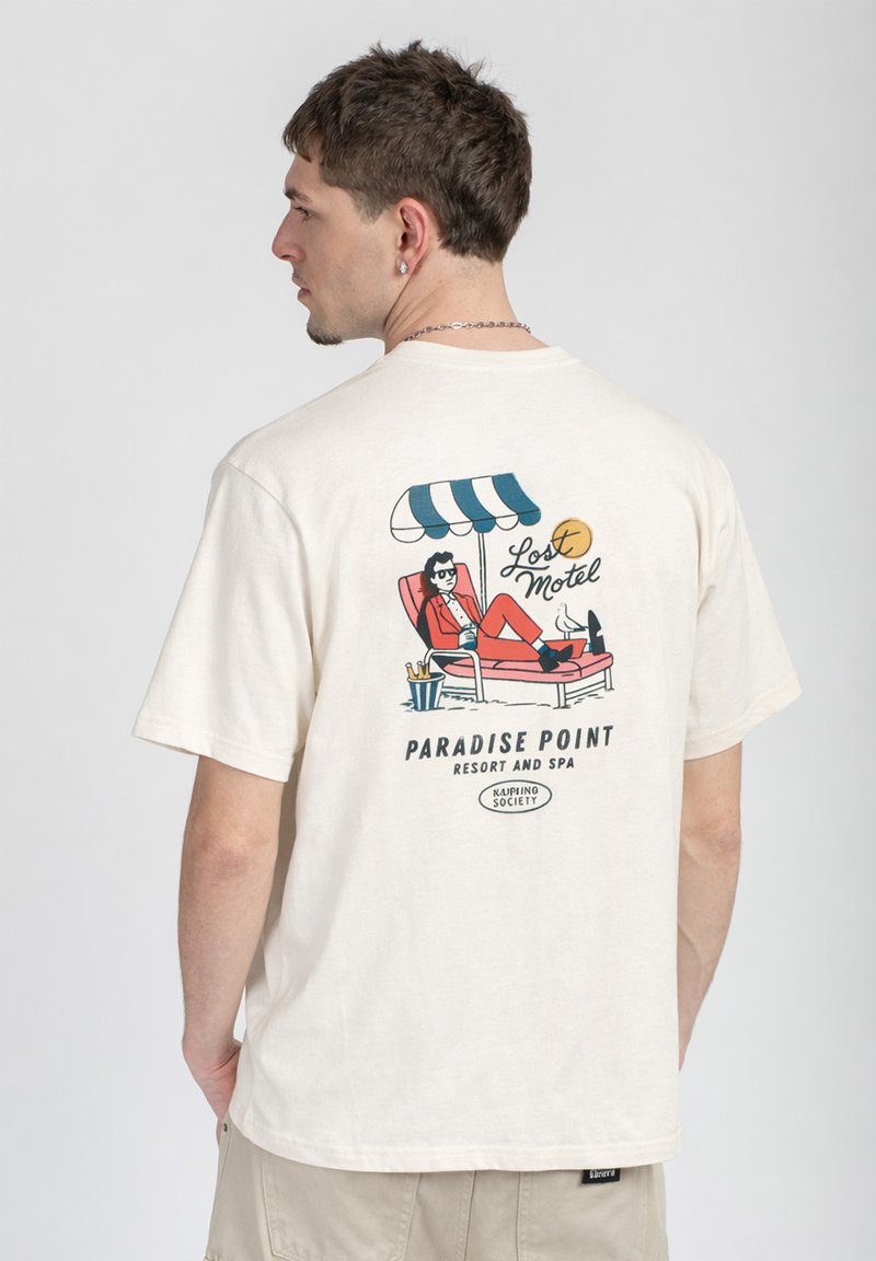Άνδρας που φοράει λευκό μπλούζα με πολύχρωμο γραφικό εικόνας ενός ατόμου που χαλαρώνει κάτω από ομπρέλα σε τυρκουάζ και λευκό, με το κείμενο "Lost Motel Paradise Point Resort and Spa."