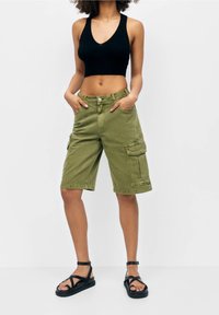 Kvinde i sort ærmeløs crop top, olivengrønne knælange cargo shorts og sorte sandaler med stropper, stående med hænderne i lommen.