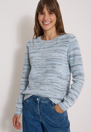 Femme souriante portant un pull en tricot bleu clair et gris à manches longues et un jean bleu avec une main dans la poche.
