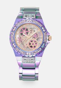 Montre multicolore avec un bracelet et un boîtier métalliques violets, un cadran transparent avec des mécanismes visibles, des accents roses et une lunette incrustée de cristaux.