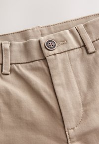 Close-up af beige bukser talje, der viser bæltestropper og en knap mærket "Authentic Next Dude."
