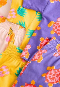 Två färgglada kläder liggande platt: en gul med ananas och blå bladmönster, och en lila med rosa sköldpaddsmotiv, båda med elastiska band.
