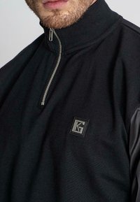 Černý pulovr s krátkým zipem a strukturovanou tkaninou, s vroubkovaným límečkem a malým embosovaným logem v obdélníkové nášivce na přední straně.