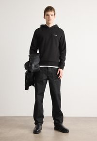 Sort hettegenser med frontlogo, brukt med vide mørke jeans. Modellen holder en svart skinnjakke og har på seg svarte støvler. Ensfarget bakgrunn.