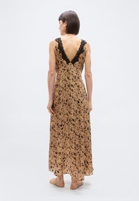 Γυναίκα που στέκεται ξυπόλητη, φορώντας ένα αμάνικο, μακρύ μέχρι τον αστράγαλο φόρεμα καφέ με μαύρα λεοπάρ στίγματα και μαύρη δαντέλα στην βαθιά V πλάτη.