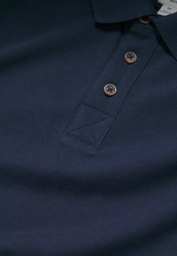 Sötét tengerészkék polo ing, texturált anyagból készült, három barna gombbal és háromszög alakú varrással a gombolósávon.