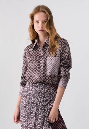Donna con capelli lunghi e mossi che indossa una camicia viola a bottoni con motivo e una gonna abbinata su uno sfondo chiaro uniforme.