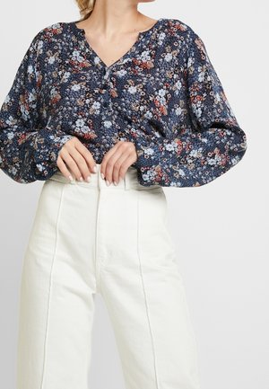 Langärmlige, marineblaue Bluse mit floralem Muster in Blau und Rot, ausgestattet mit einem knöpfbaren Ausschnitt und weichem Stoff. Kombiniert mit hoch taillierten weißen Hosen.