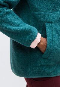 Τζάκετ από φλις σε τιρκουάζ χρώμα με απαλό υφή και ροζ βολάν στο μανίκι, με πλαϊνή τσέπη και λεπτομέρειες από λείο ύφασμα.