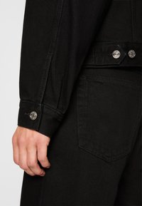 Black denim jacket with a textured finish, featuring silver hardware, buttoned cuffs, and a visible pocket.