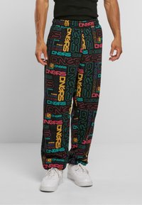 Nedre del av en person som har på seg flerfargede joggebukser med "DNGRS"-tekstmønster og hvite sneakers, stående mot en ensfarget bakgrunn.