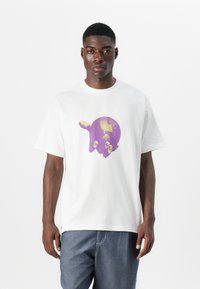 T-shirt en coton blanc avec un graphisme de main violette tenant un globe. Manches courtes et coupe décontractée avec un col rond.