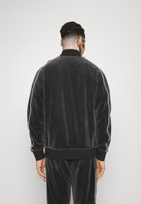 Černá sametová bomber bunda s límcem stojáčkem, bílými bočními pruhy, elastickými manžetami a pasem. Hladká textura, minimalistický design.