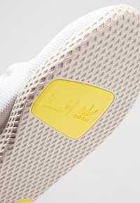 Sula i vit gummi med ett texturerat mönster. Har en gul etikett med "adidas"-logotypen och texten "Pharrell Williams".