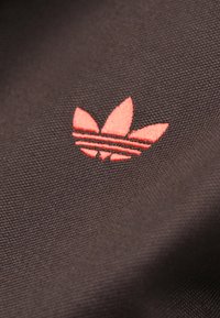 Must kangas tekstuurse pinnaga, kus on punase tikandiga Adidas logo, millel on kolm lehekaunistust ja all kaks horisontaalset triipu.