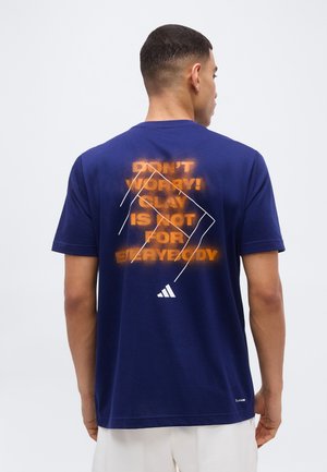 Homme de dos portant un t-shirt bleu marine avec un texte orange "NE VOUS INQUIÉTEZ PAS ! LE JEU N'EST PAS POUR TOUT LE MONDE" et des lignes géométriques blanches au dos.