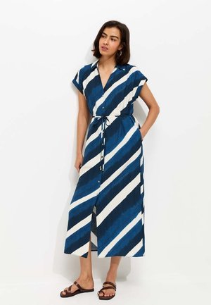 Kobieta w niebiesko-białej sukience midi w ukośne paski na guziki, z krótkimi rękawami, wiązaną w talii, w brązowych płaskich sandałach, stojąca na tle białej ściany.
