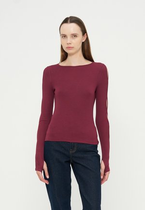 Femme aux longs cheveux bruns portant un haut bordeaux à manches longues avec des découpes aux épaules et un jean bleu foncé, debout devant un fond blanc uni.
