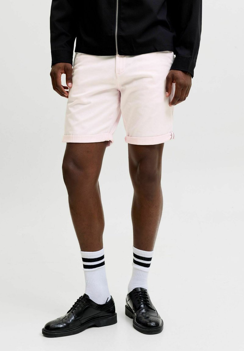 Homme portant un short rose clair à ourlet roulé, des chaussettes blanches à rayures noires, des chaussures habillées noires et une veste noire zippée sur un fond uni.