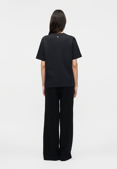 Άτομο με μακριά σκούρα μαλλιά, γυρισμένο με την πλάτη, φοράει απλό μαύρο μπλουζάκι και μαύρο παντελόνι με φαρδιά πόδια, σε λευκό φόντο.