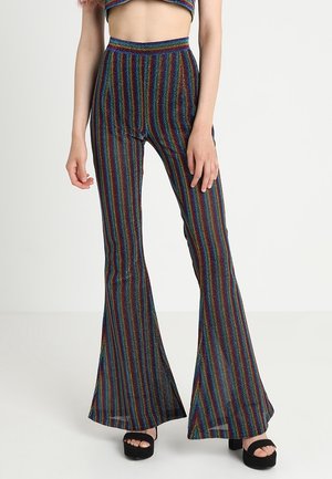 Woman wearing high-waist, rainbow-striped bell-bottom pants with a matching crop top and black platform sandals, standing against a white background.