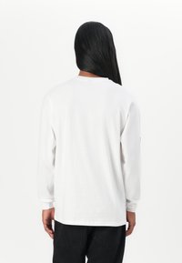White long-sleeve shirt in cotton fabric, features a round neck and loose fit, with ribbed cuffs and a small logo on the sleeve.