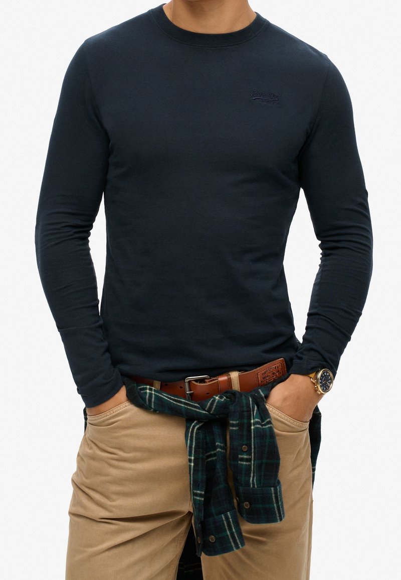 Dunkelblaues Langarm-T-Shirt mit Rundhalsausschnitt, kombiniert mit beigen Hosen und einem gebundenen grün-karierten Hemd um die Taille. Mit einem Logo auf der Brust.