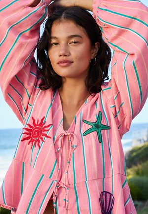 Young woman wearing loose pink striped top with sun, starfish, and shell patches, posing with arms raised near a beach setting.