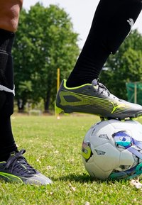 Black soccer cleats with neon accents, positioned on a patterned white soccer ball, on grassy outdoor field.