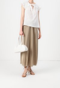 Weißes Blusenshirt mit kurzen, gerafften Ärmeln und einem gebundenen Kragen, kombiniert mit einem langen, beigen Faltenrock. Hält eine kleine weiße Handtasche in runder Form.