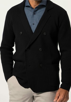 Hombre vestido con blazer cruzado negro sobre camisa azul con botones y pantalones beige, una mano en el bolsillo, torso y brazos visibles.